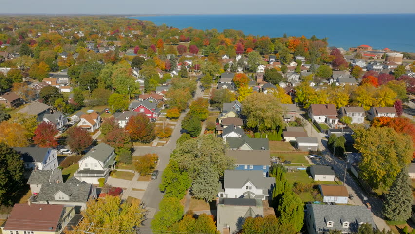 Drone aerial pulling back over Sheboygan, Wisconsin neighborhood streets and homes surrounded by peak autumn color with Lake Michigan on the horizon under a clear blue sky.