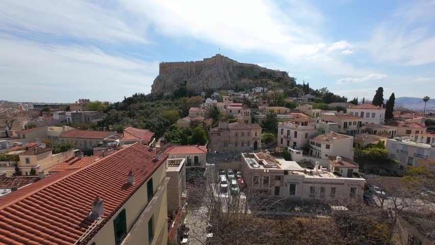 Cinematic FPV drone shot of traditional Byzantine church in the Plaka district of Athens, Greece. Beautiful view of red rooftops, narrow streets, and the Acropolis in the background.