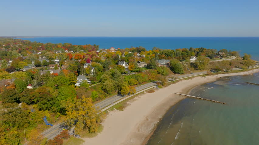 Drone aerial over Lake Michigan toward the Sheboygan, Wisconsin shoreline and road with charming neighborhood homes and colorful autumn trees beneath a clear blue sky.