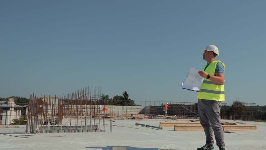 Architect reviewing blueprints at modern construction site. An architect in a reflective vest and helmet examines building blueprints while walking through a large open construction site.