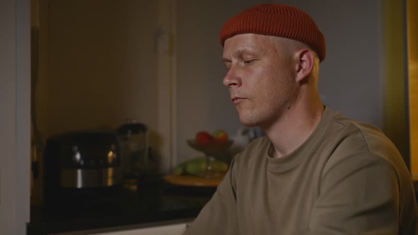 Side view of a man eating chips in the evening kitchen. Warm light and relaxed posture emphasize the calm and mindful atmosphere of his ritual. High quality 4k footage