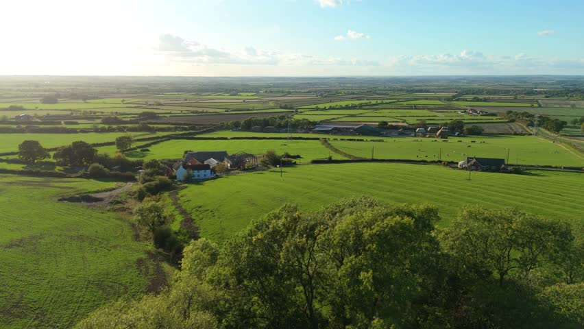 Aerial view of lush green fields interspersed with farmhouses and barns, creating a patchwork of textures and tones under a clear blue sky, Melton Mowbray, England, United Kingdom.