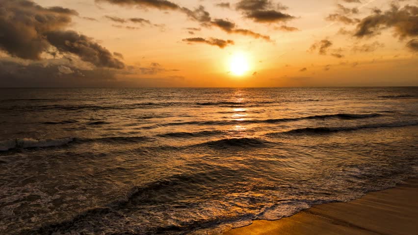 Aerial view of a serene ocean reflecting the golden sunset light over the horizon, as waves gently meet the sandy shore, Waikkal, North Western Province, Sri Lanka.