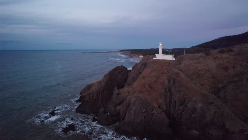 A white lighthouse overlooks the coastline of Japan, offering a serene coastal view as daylight fades.