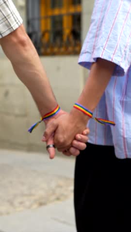 Romantic close up of two men holding hands while walking down a city street, proudly wearing rainbow bracelets symbolizing love, equality, and lgbtq plus community pride during a sunny day