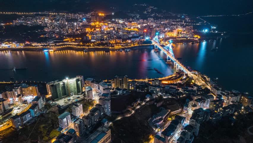 Stunning aerial night view of Badong city in Enshi, Hubei Province, China showcasing illuminated bridge over river with colorful city lights reflected in water below.