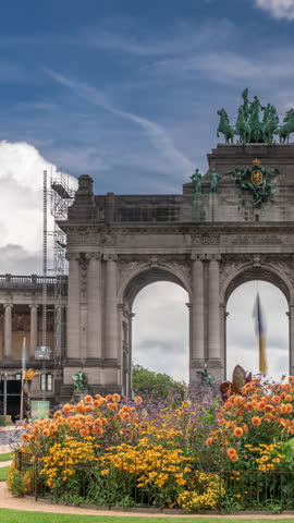 Timelapse of the Cinquantenaire Arcade in Jubelpark with flowerbed, Brussels, Belgium. The memorial triumphal arch stands tall under dramatic clouds, symbolizing Belgian history and independence.