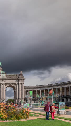 Timelapse of the Cinquantenaire Arcade in Jubelpark, Brussels, Belgium. The memorial triumphal arch with flowerbed stands tall under dramatic clouds, symbolizing Belgian history and independence.