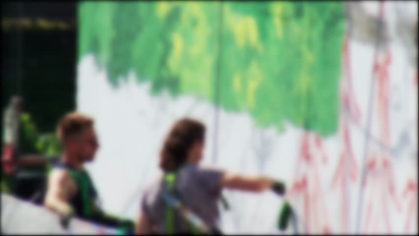Wide-Angle Blurred View Of Artists Painting Large Building Mural. Defocused Scene Shows Two People On Lift Creating Colorful Street Art.