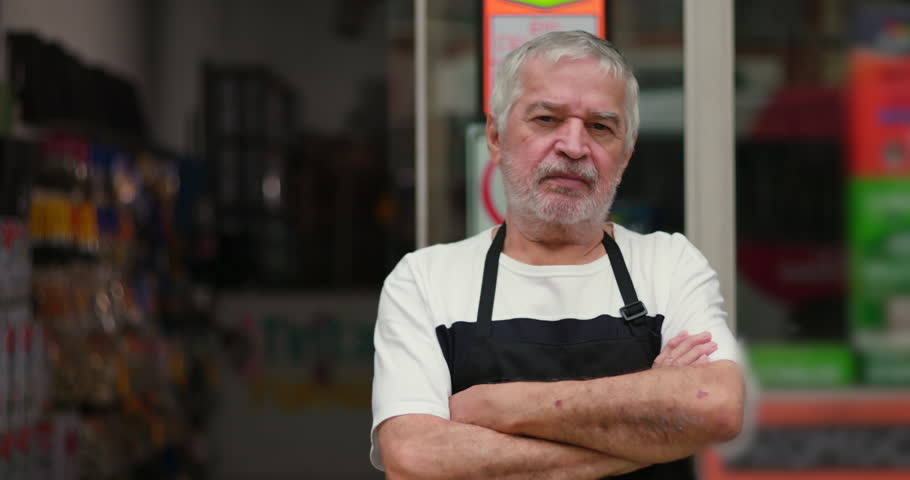 Elderly man with folded arms standing outside store, wearing apron, looking serious and confident, urban retail setting, storefront background