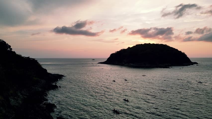 Stunning sunset view over Yanui Beach in Phuket, Thailand, with small boats scattered across calm sea and silhouetted island in golden light.