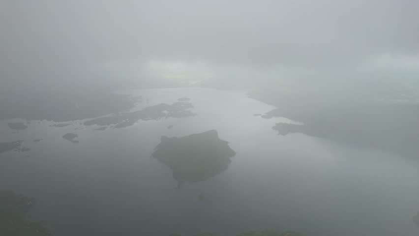 An aerial shot of lake with several small, forested islands, with a thick layer of clouds and mist obscuring the sun and casting shadows on the water.
