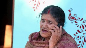 A senior Indian village woman sits outside her house with a blue wall, talking on her cellphone. The warm light and rustic setting reflect rural connection, culture, and simple daily life. - Powered by Shutterstock - Get 15% off with code: PIKWIZARD15