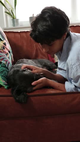 Young girl tenderly cuddling and kissing her small black dog while relaxing on the sofa at home