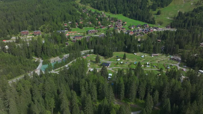 Beautiful aerial drone footage of La Fouly, a picturesque mountain village in the Swiss Alps. The video shows green forests, alpine meadows, and cozy wooden chalets under bright summer sunlight. 