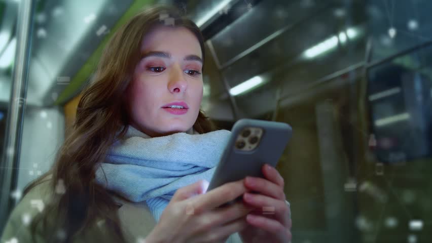 Caucasian woman wearing light scarf. Holding smartphone and reading messages while riding subway at night. Female looking at screen as glowing chat bubbles appear in air above phone. Public transport.