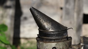 Aged bee smoker emitting smoke amidst beekeeping tools and wooden hives under bright sunlight, showcasing traditional apiculture equipment and practices in a rural setting. - Powered by Shutterstock - Get 15% off with code: PIKWIZARD15