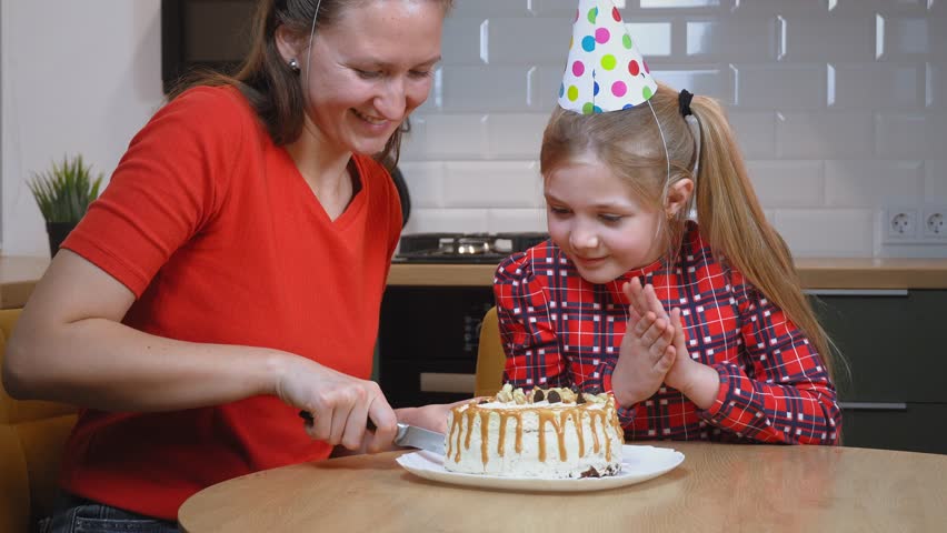 Mother in red t-shirt and party hat joyfully cutting birthday cake alongside daughter wearing plaid shirt, sharing loving moment in family kitchen during festive celebration