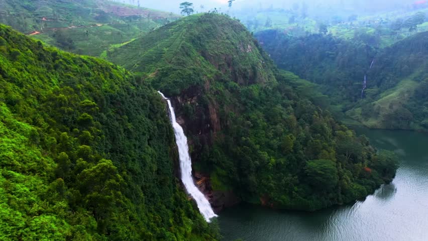 A breathtaking aerial view of a tall waterfall cascading down a lush green hillside into a tranquil lake, surrounded by misty mountains and dense tropical forest. - Powered by Shutterstock - Get 15% off with code: PIKWIZARD15