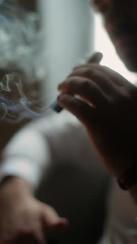 Close-up of man smoking cigar in dim light, stylish male hand with wristwatch holding cigar, smoke rising softly in cinematic atmosphere, concept of luxury, relaxation, and masculinity