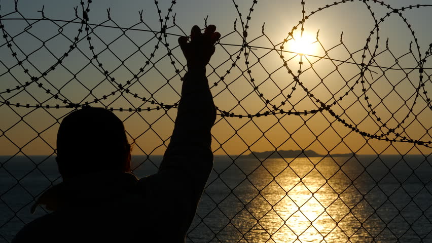 Prisoner touching barbed wire fence at sunset. Silhouetted prisoner reaching toward barbed wire fence at golden sunset, symbolizing deep longing for freedom and hope beyond confinement's boundaries