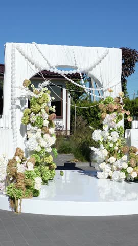 Elegant wedding arch on the podium with fabric, beads, candles, tiers and romantic floral arrangement in white and green tones. Amaranth, anthurium, hydrangea, rose, dahlia. Summer windy atmosphere.