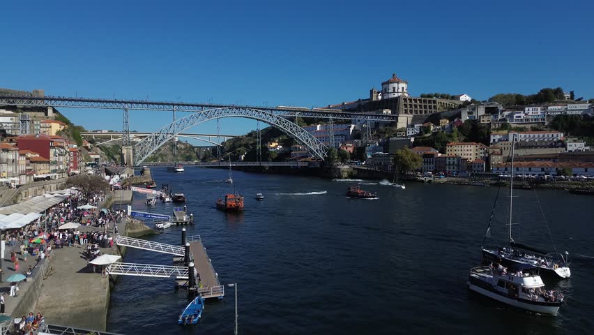 Aerial view of Porto, Portugal, showing the Dom Luís I Bridge, red-roofed historic buildings, and the Douro River. People walk across the bridge, and boats line the waterfront under clear blue skies.