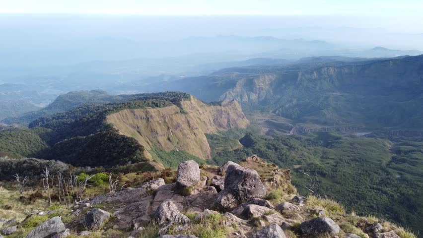 A 14-second aerial drone footage showcasing a breathtaking view from a mountain peak overlooking vast green valleys and steep hills. Large rocks in the foreground emphasize the raw and natural beauty 