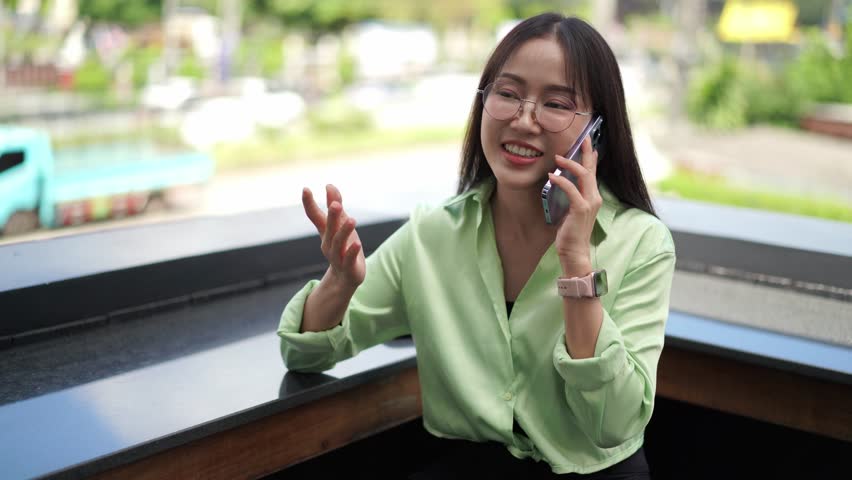 Smiling young asian woman with glasses having an enjoyable conversation on her smartphone outdoors
