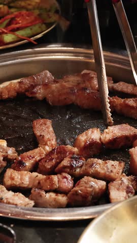 Pork belly pieces grilled on tabletop barbecue, tongs turning meat, warm lighting, close-up view