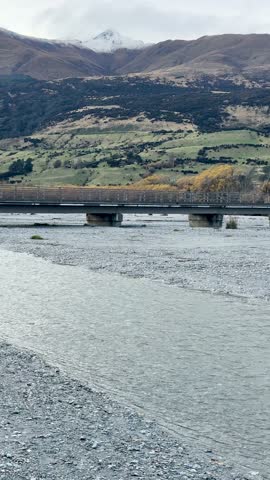 Camera slowly pans across river, bridge, and mountains under overcast daylight in Glenorchy, New Zealand