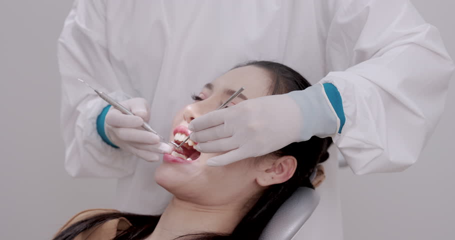 Asian female patient lying on dental chair receiving oral examination from dentist using mouth mirror and sickle probe while wearing white coat and latex gloves in clinical setting