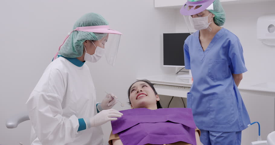 Asian woman undergoing light cured dental restoration using orange curing shield while dentist applies resin with explorer tool in clinical setup with assistant monitoring progress