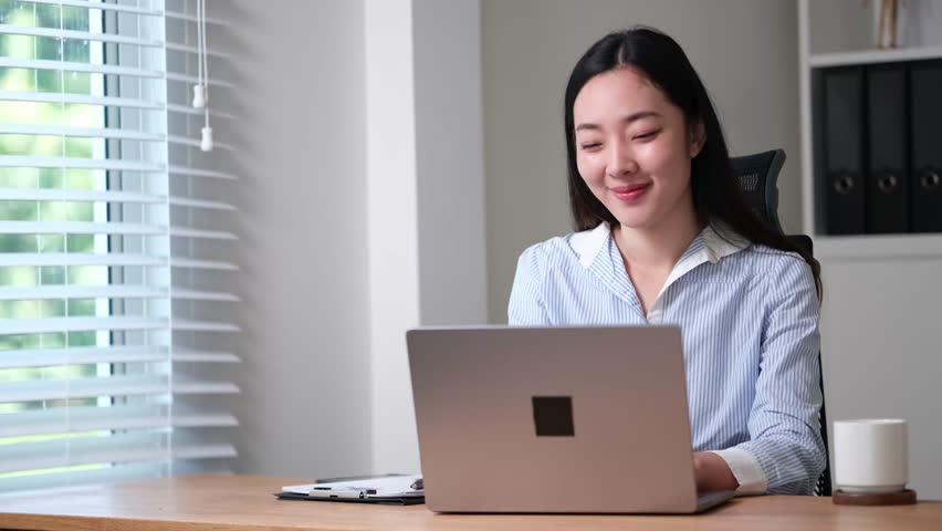 Smiling young woman working on a laptop in a modern office, sitting at a desk with natural light from window blinds.