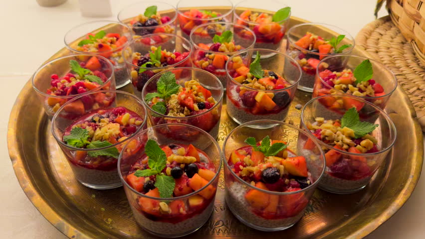 Close-up of several glass cups filled with chia pudding, fresh fruit, nuts, and mint leaves arranged on a golden tray under warm light.