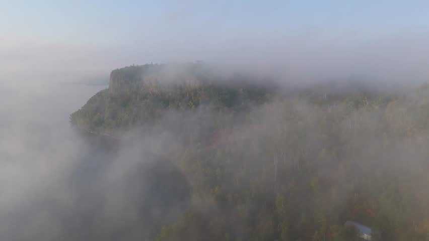 Dense morning fog drifts over the rugged shoreline of Lake Superior, revealing dramatic vertical cliffs and early autumn forest hues in Ontario, Canada. Aerial view