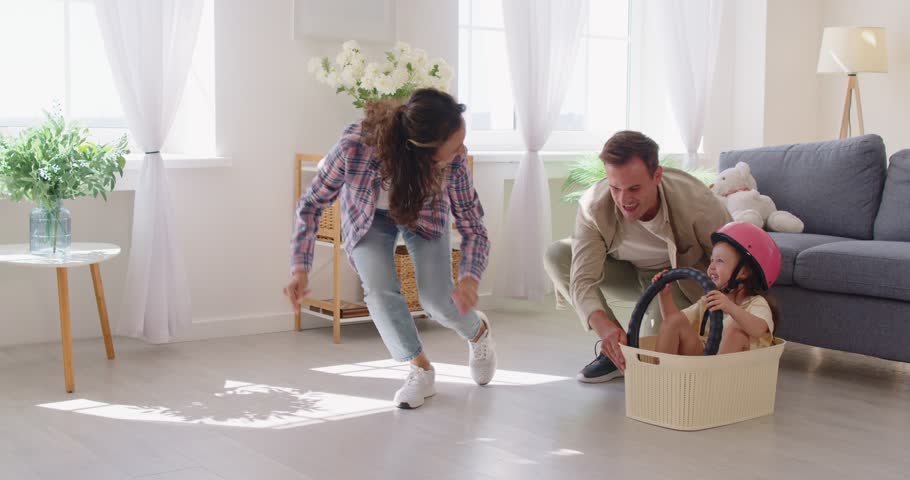 Family at home, daughter play driving in box car. Dad pushes a basket across living room as Mom claps the happy child girl grips a steering wheel and wears a helmet. Joyful parents having fun.