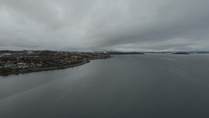 Aerial drone footage capturing a Norwegian fjord under gloomy evening weather. The scene features calm, dark waters surrounded by misty mountains and low clouds, creating a moody and atmospheric view 