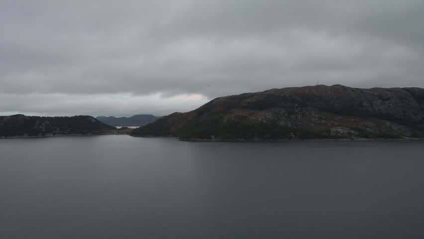 Aerial drone footage capturing a Norwegian fjord under gloomy evening weather. The scene features calm, dark waters surrounded by misty mountains and low clouds, creating a moody and atmospheric view 