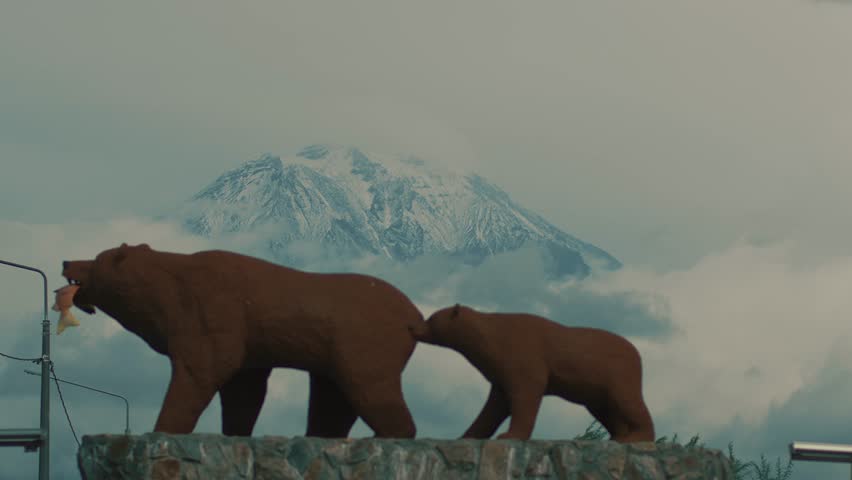 Famous sculpture of she-bear with fish in mouth and bear cub on pedestal at exit of city of Yelizovo against backdrop of grandiose snow-capped volcano