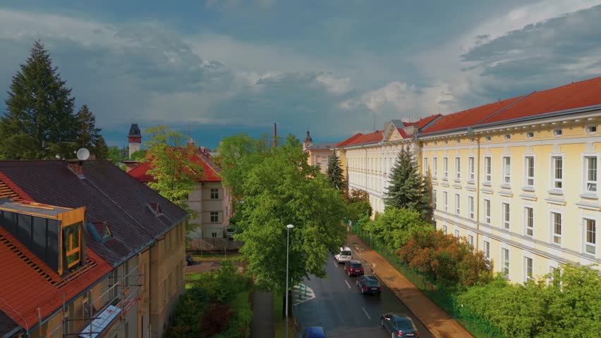 aerial perspective showcasing the charming cityscape of České Budějovice, Czech Republic, under a dramatic, cloudy sky. This footage captures the unique architecture, vibrant rooftops, and lush green