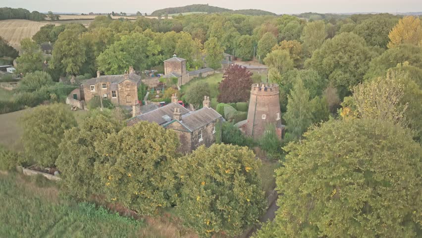Ascending Aerial view of the round crenellated tower and adjacent stone house in Wentworth Castle Gardens, South Yorkshire, England, surrounded by autumn trees and historic parkland