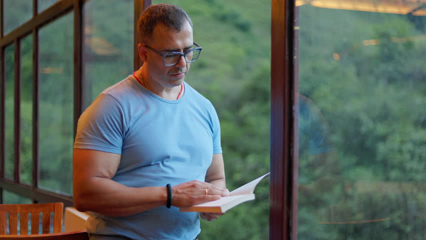 man relaxing and reading a book indoors during leisure time with greenery view.