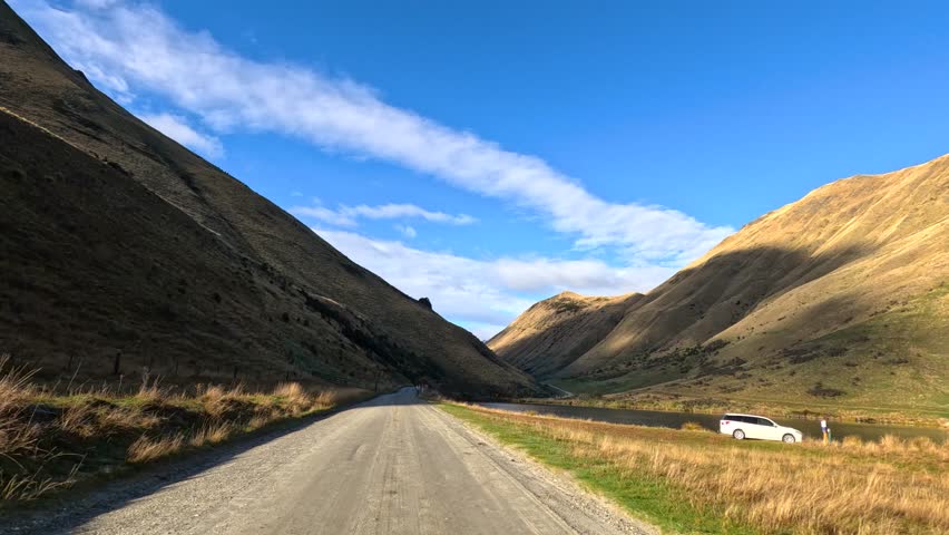 Vehicle drives on gravel road between sunlit hills under clear blue sky, camera moves forward.