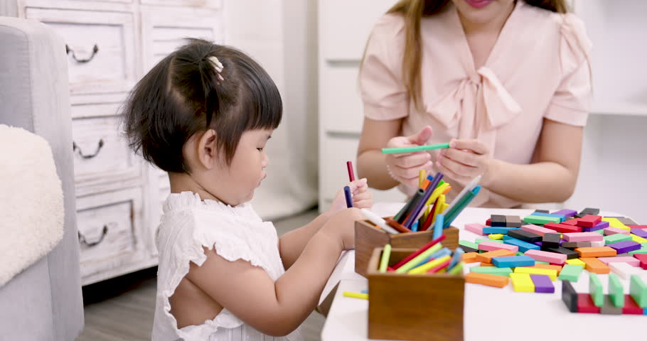 young asian preschool girl exploring colorful pencils with curiosity while seated at table as asian female teacher guides her through early education learning activity indoors