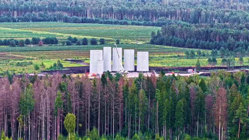 Aerial view of cranes assembling large silos or windmills amid forest and open nature