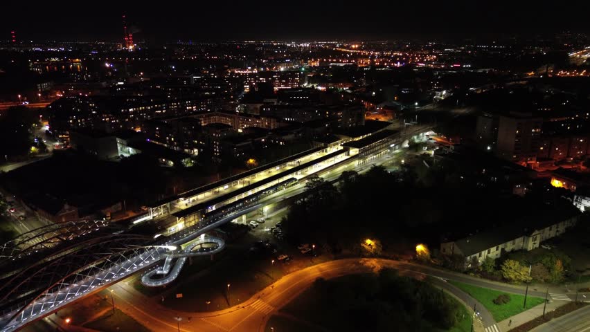 Aerial view of a train station at night, showcasing illuminated platforms and surrounding cityscape, as the camera smoothly pans to reveal the vibrant urban environment