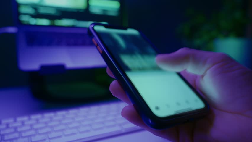 Hand holding smartphone and quickly scrolling social media feed in dark room with desk on background, illuminated by blue and purple neon light. Digital addiction and mindless content consuming