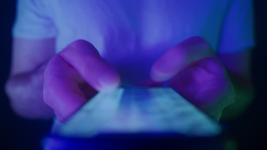Close-up of hands holding a smartphone and quickly scrolling through social media feeds at night in neon light. Doom scrolling, phone overuse and digital addiction concept. 