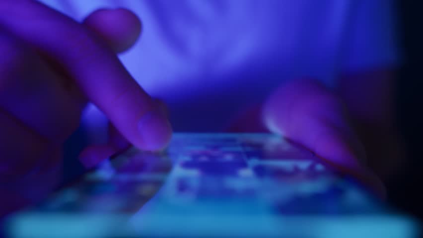 Close up of hands using a mobile phone in low light, browsing social networks and consuming endless online content. Doomscrolling and information overload.